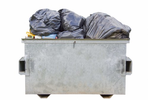 A large, industrial-style metal skip situated on a paved surface, filled to capacity with black heavy-duty garbage bags made of thick plastic, some of which are partially crumpled or slightly opened, revealing an assortment of waste materials inside. The skip has a rectangular shape with smooth, painted metal sides and reinforced edges, positioned outdoors with no visible background, suggesting a commercial or residential rubbish removal setting, possibly in Cricklewood. The scene is lit with natural daylight, emphasizing the metallic texture of the skip and the glossy finish of the waste bags. This image vividly depicts a typical waste collection scene that might be encountered when using Commercial Waste Removal Cricklewood for rubbish disposal services, showcasing the type of waste and containers involved in professional rubbish clearance in that area.