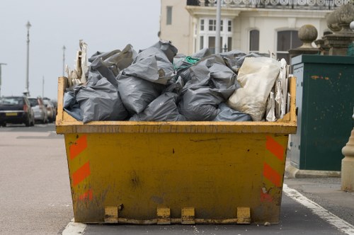 A large yellow plastic skip filled with numerous black and white rubbish bags, situated on a paved surface in an urban setting. The bags appear to contain general waste and are tightly packed. The skip's surface shows signs of dirt and wear, with orange and red reflective safety markings on its sides. In the background, there are parked cars, a building with a light-colored facade, and a row of rooftops, indicating a residential or commercial area possibly in Cricklewood or nearby northwest London. The environment is outdoors with natural daylight filling the scene, and the waste removal service provided by Commercial Waste Removal Cricklewood caters to local businesses or residents needing rubbish clearance services in this area.
