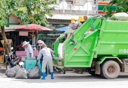 A bright green commercial waste collection truck with a large, open rear compartment is parked on a street, while two workers in protective clothing, including white suits and blue rubber boots, are placing black rubbish bags into the truck's loading area. The scene takes place in an urban environment with trees, small shops, and market stalls visible in the background, along with some colorful signage and awnings. The workers are actively engaged in waste collection, and the street appears clean and organized, reflecting typical rubbish removal operations in a residential or small commercial area possibly within Cricklewood, North West London. The truck, part of a professional waste management service, displays company branding characteristic of local rubbish removal services in the UK, supporting the context of commercial waste disposal.