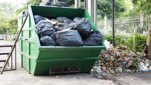 A large green metal skip filled with black plastic rubbish bags, situated on a paved area outdoors. The bags appear to contain general waste and are piled up to the top of the container. To the right of the skip, there is a small pile of loose rubbish, including paper, plastic, and organic debris, scattered on the ground near a low fence and some trees. The background features a metal fence, greenery, and a park-like setting, indicating the waste is temporarily stored outside for collection. This scene reflects typical rubbish removal activity, relevant to commercial waste services offered by Commercial Waste Removal Cricklewood, serving local areas and businesses in the Cricklewood postcode district.