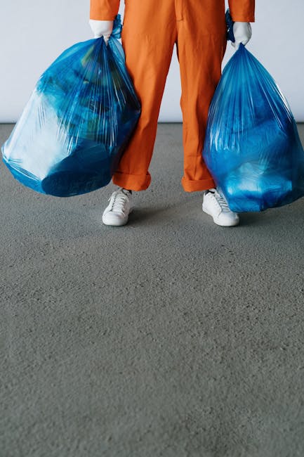 A person standing on a grey indoor surface holding two large blue plastic rubbish bags, one in each hand. The individual is wearing orange overalls and white sneakers, with their legs slightly apart. 